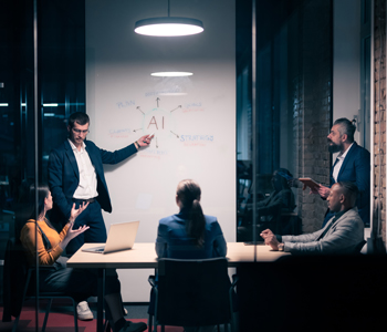 Diverse board meeting with employees sitting at table late at night. Leader points to large whiteboard that has AI written on it.