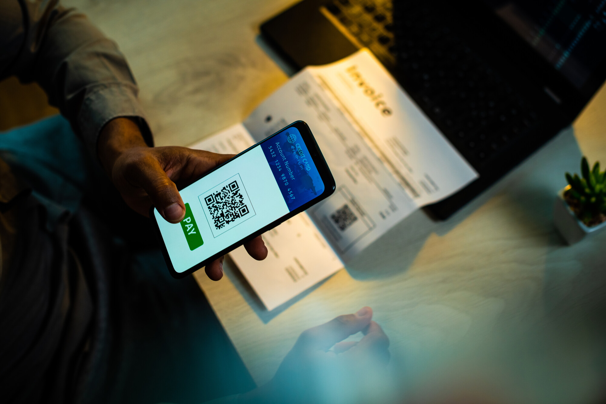 Hand holding cellphone scanning a QR code on a paper invoice laying on table in order to pay digital credit card.