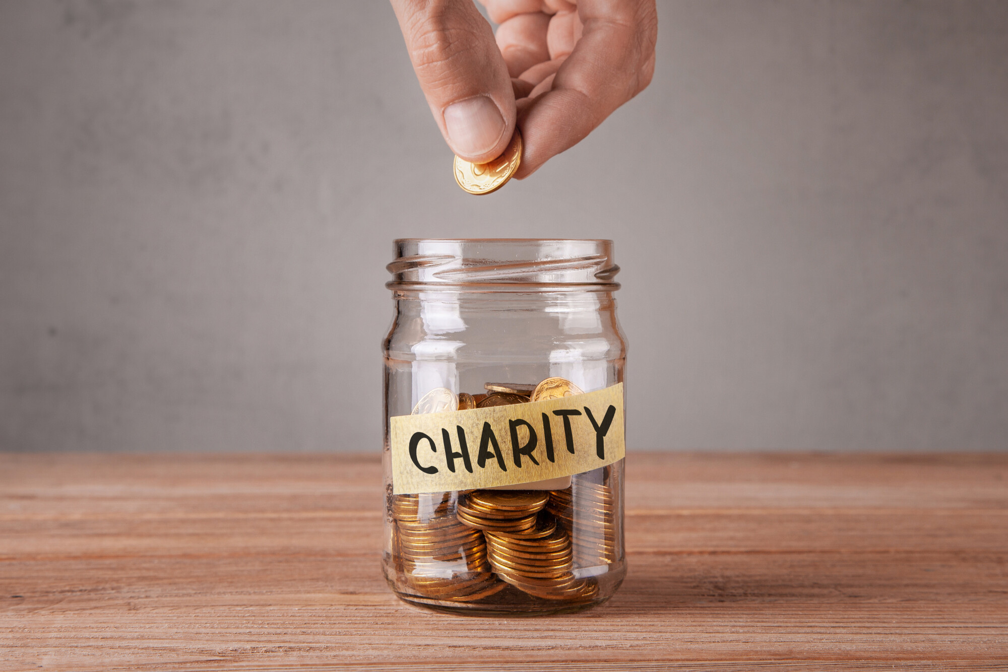 Hand hovers above ready to drop coin in glass jar full of coins with piece of tape that says Charity. 