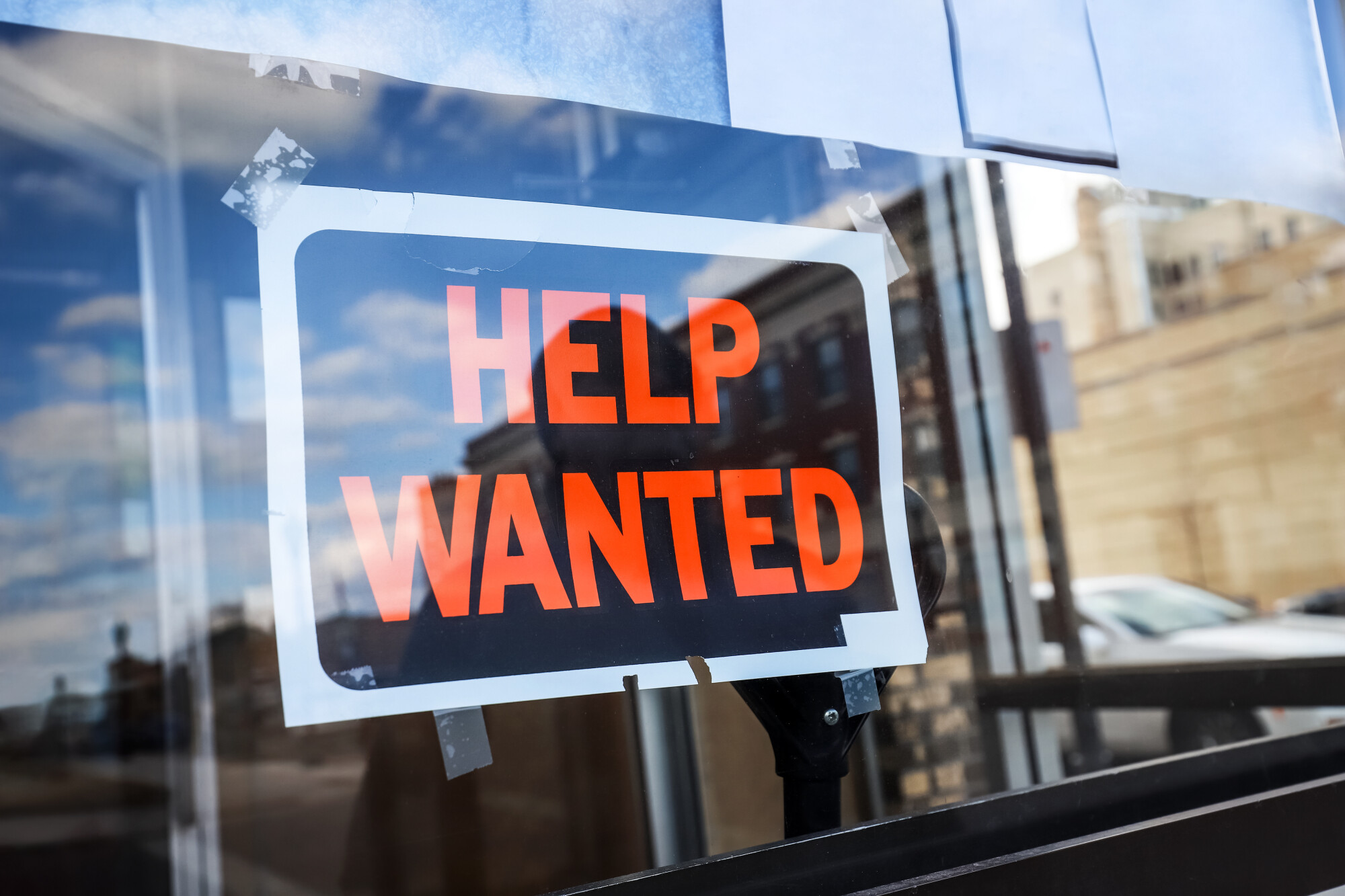 Black sign with orange letters saying Help Wanted in reflecting window of store.