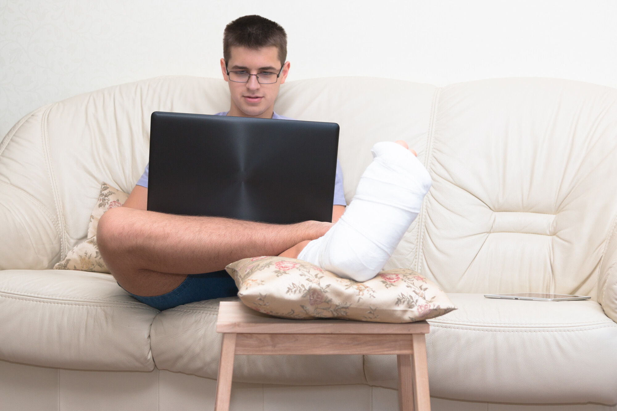 Teenage man wearing glasses sits on couch with a laptop in his lap and his injured foot wrapped in bandages and propped up on pillow on a stool.