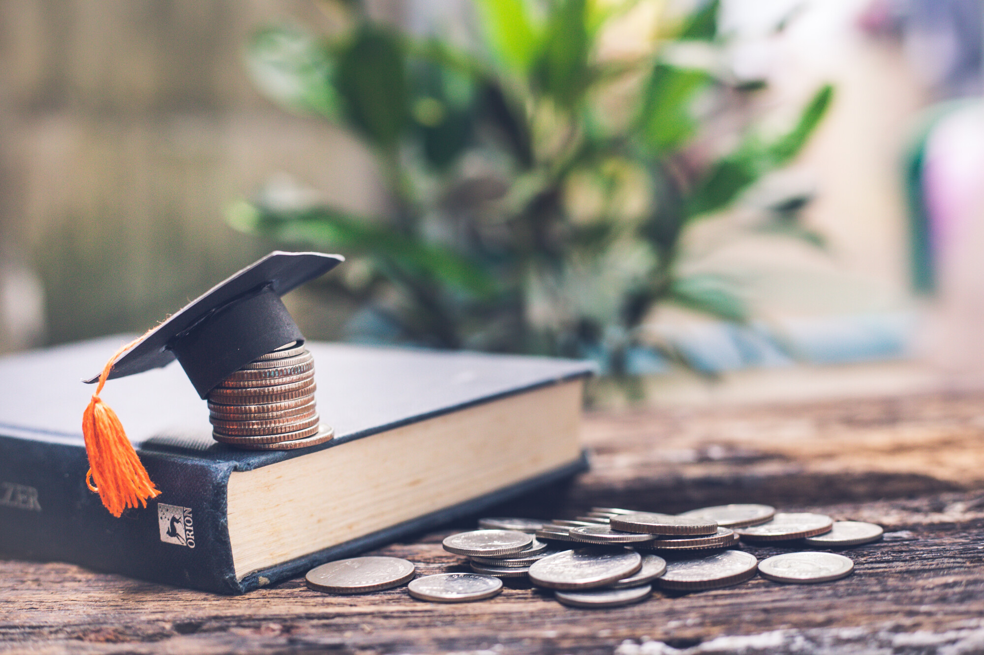 Book with miniature black graduation cap on top of stack of coins while book lays on desk next to stack of coins