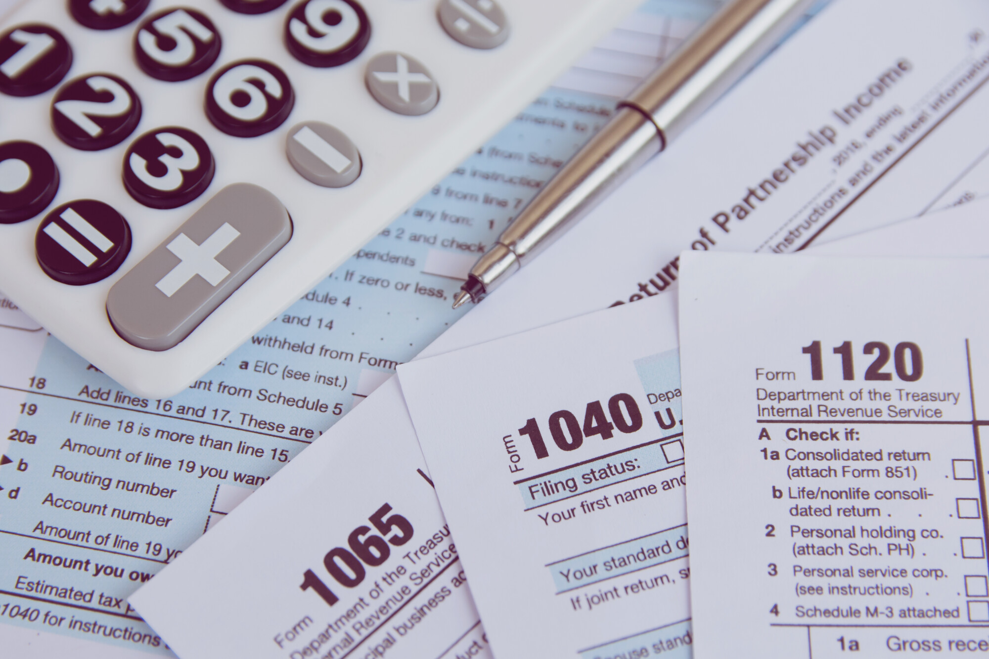 Various tax forms stacked on top of each other with calculator and pen above them.