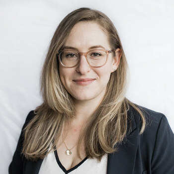 Sacha Markano-Stark is a white woman in her 30s with long blonde hair and glasses with rose-colored frames. She is wearing a white shirt with black trim, a black suit jacket, and a necklace, and is smiling.