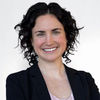 Brenna Kearns is a white woman in her 30s with brown hair. She is wearing a dark suit and metallic earrings and is smiling.