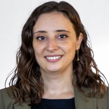 Megan Russo is a white woman in her thirties with wavy brown hair. She is wearing a white blouse and a patterned blazer and is smiling.