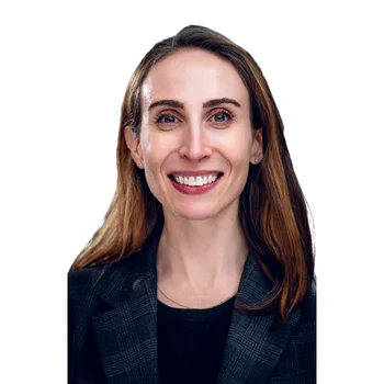 Lila is a white woman in her mid-30s with brown hair and eyes. She is wearing a black blazer and is smiling.